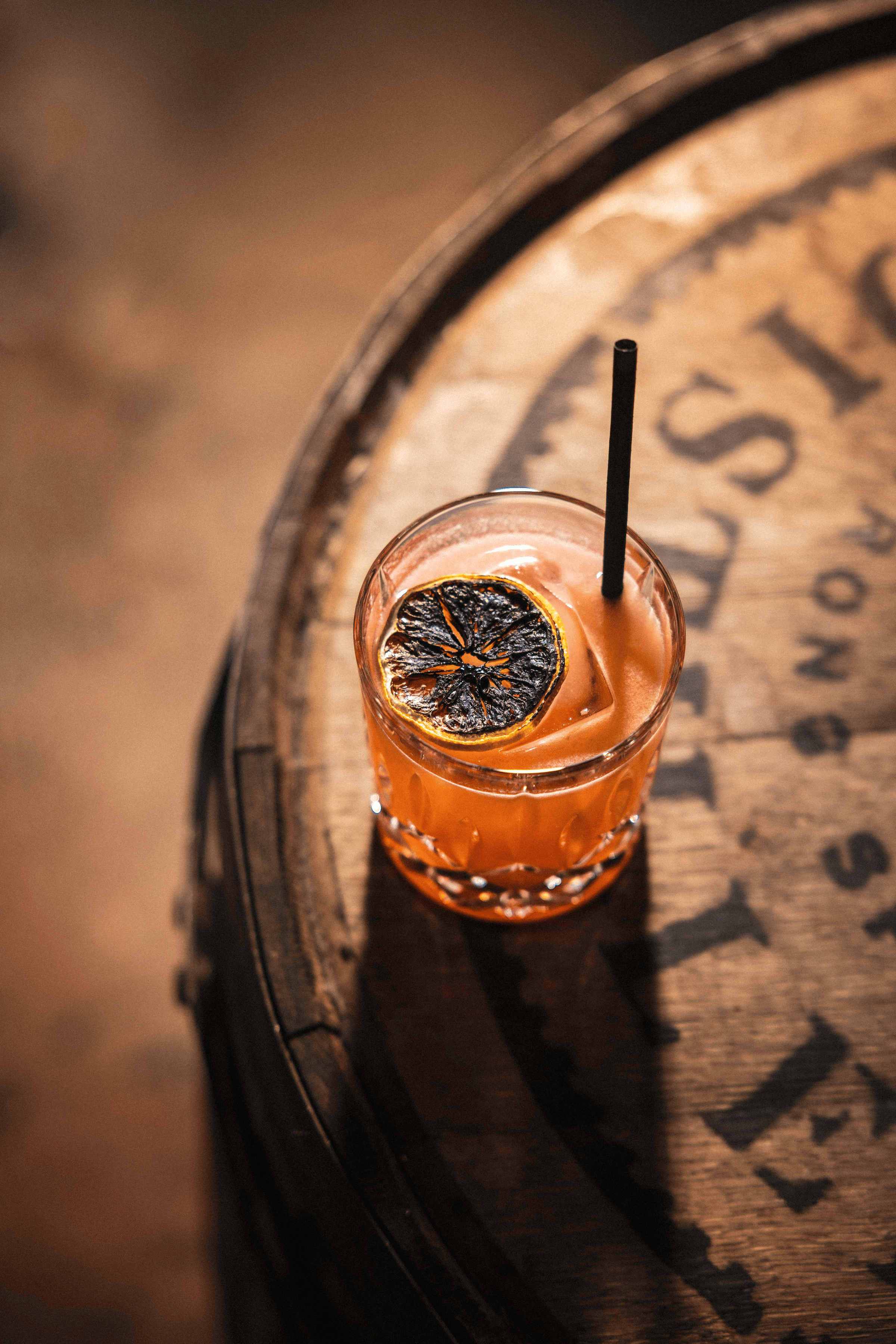 A vibrant orange cocktail rests on a dark wooden barrel, garnished with a charred lemon slice and a black straw.