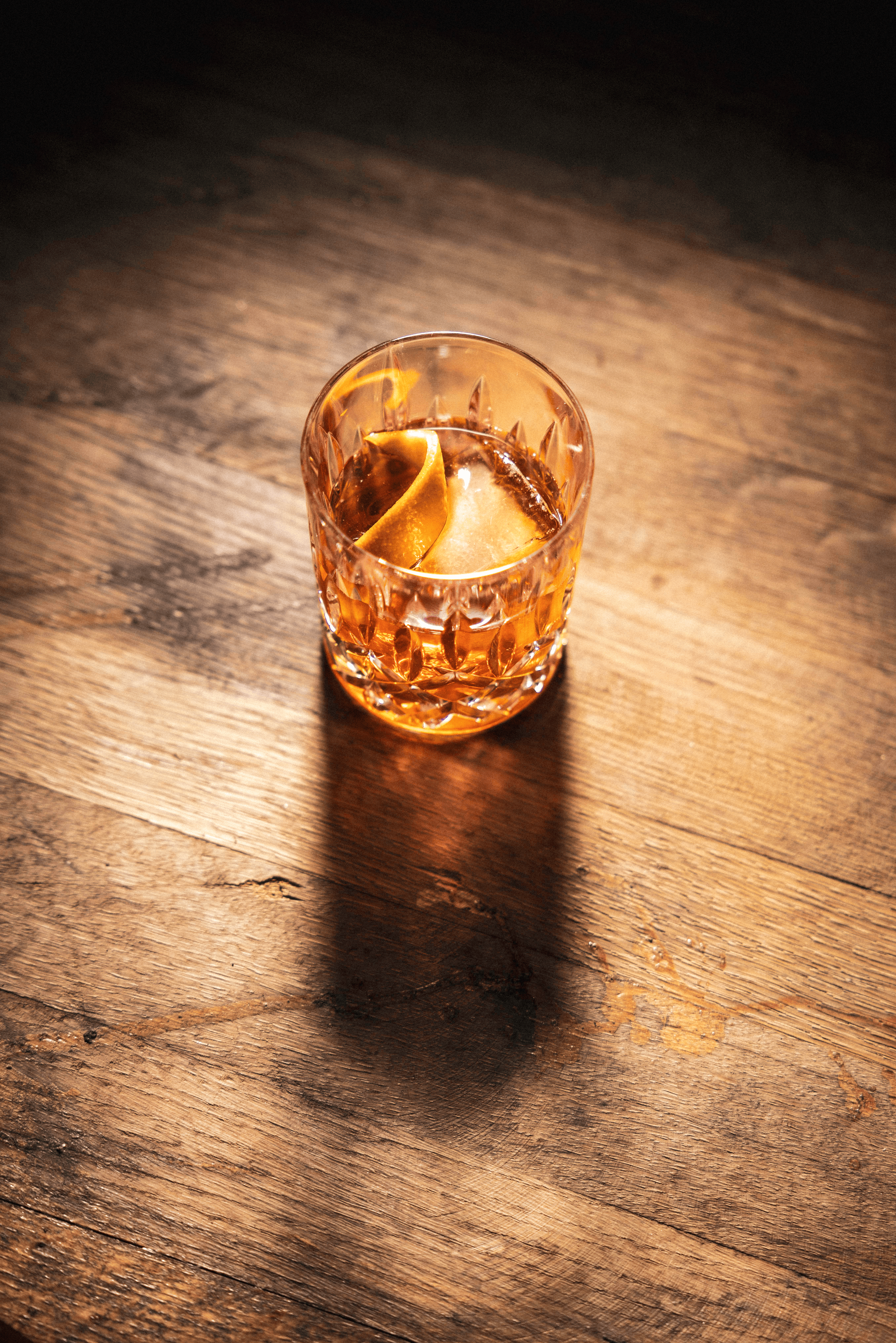 A glimmering glass of amber cocktail rests on a rustic wooden table, accented by a twist of citrus atop ice.