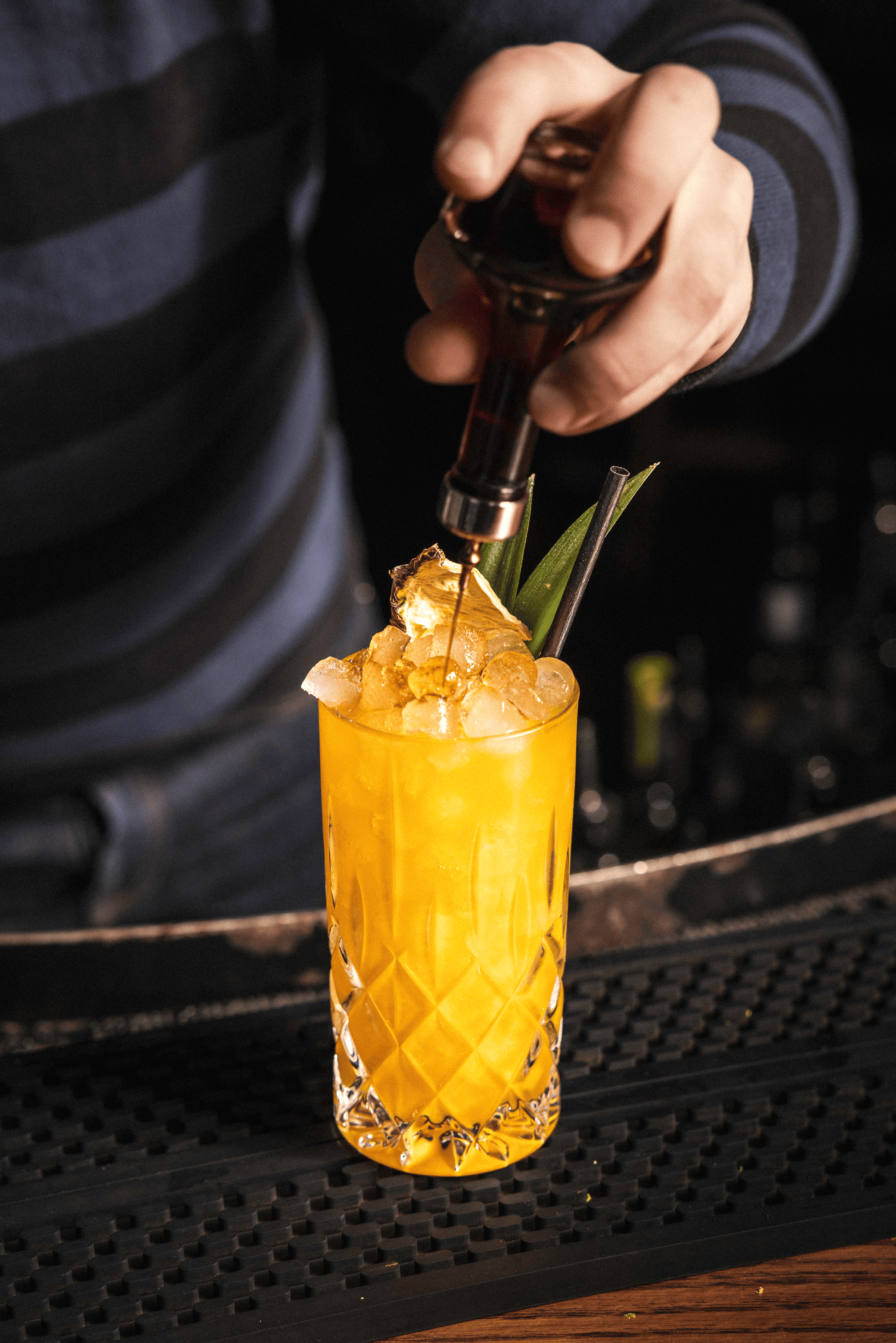 A bartender carefully drizzles a golden syrup over crushed ice in a beautifully crafted glass filled with vibrant yellow cocktail.