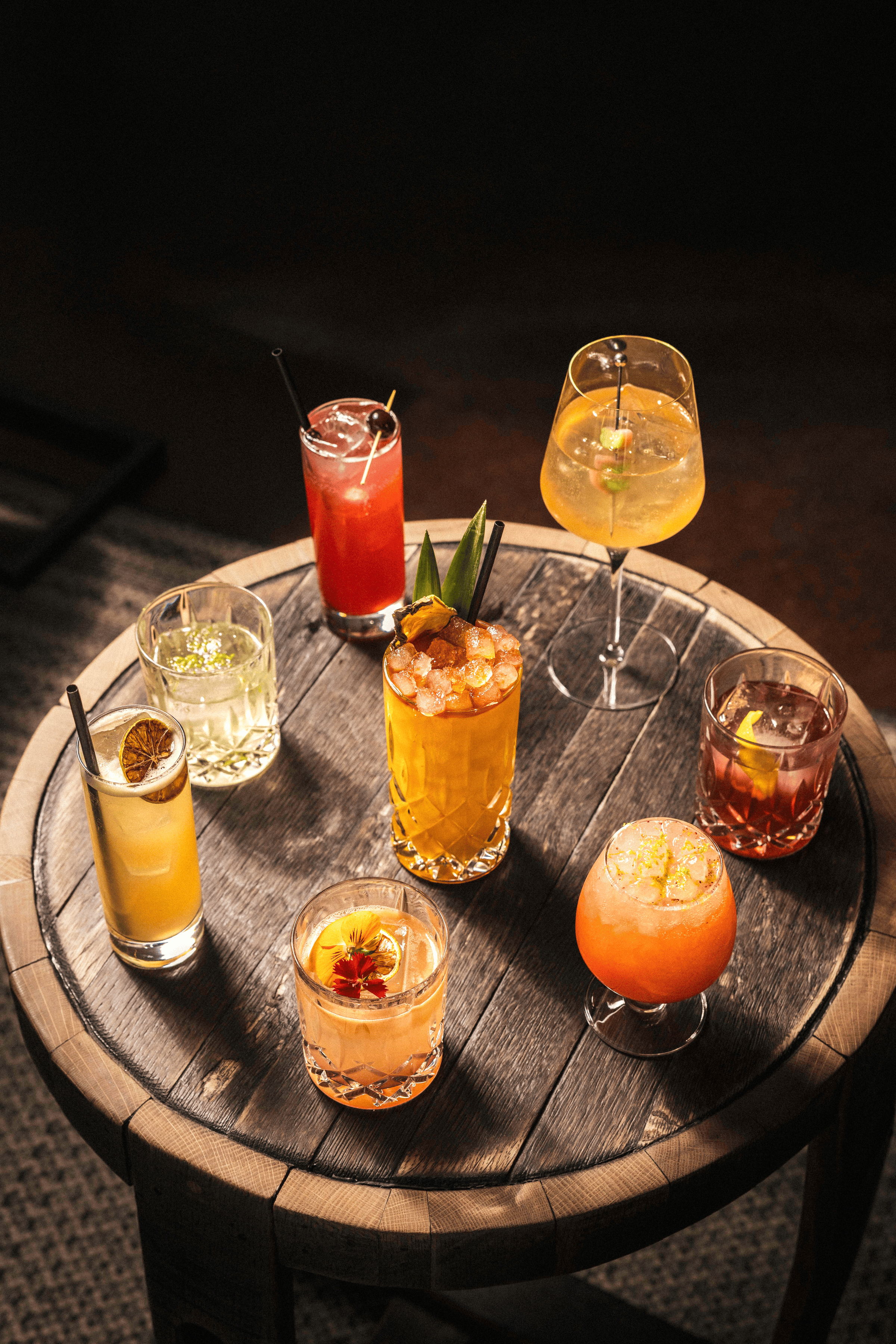 A vibrant array of colorful craft cocktails rests on a rustic wooden table, illuminated by warm, ambient lighting.
