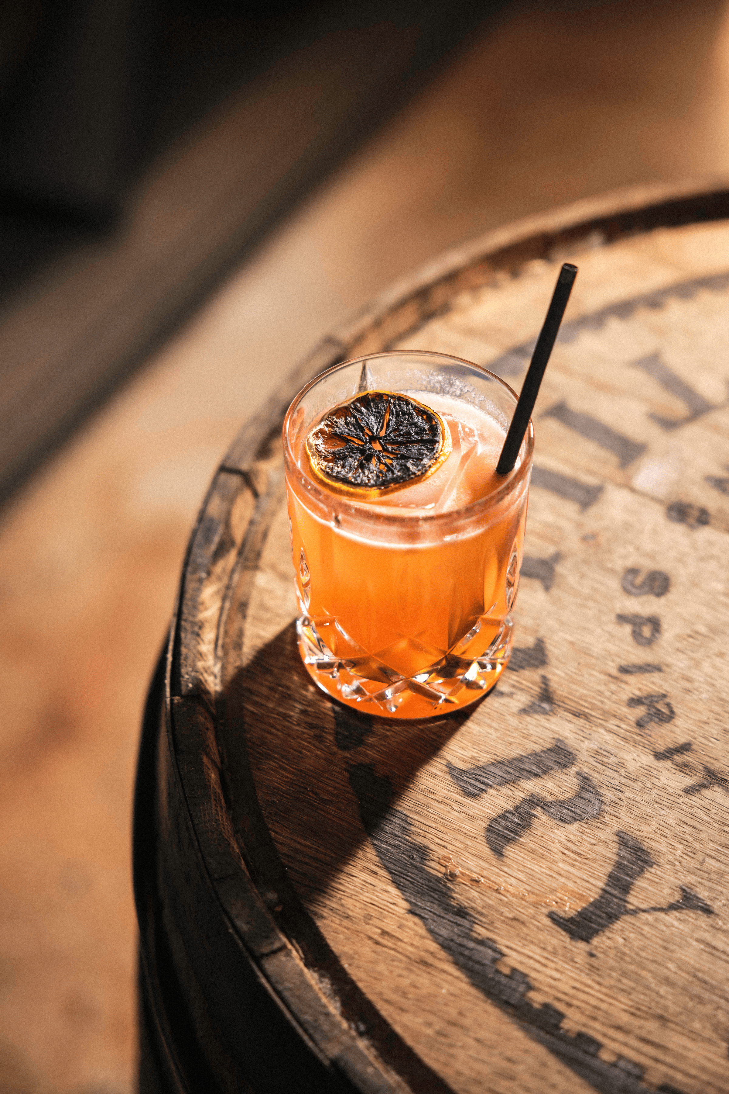 A crystal glass holds a vibrant orange cocktail, garnished with a dark, dried citrus slice, resting atop a wooden barrel.